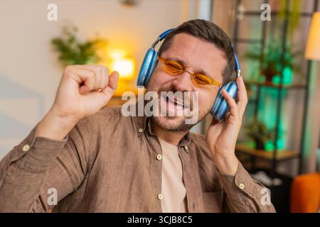 Giovane uomo d'affari felice e rilassato in ufficio che indossa cuffie e occhiali da sole ascoltando la musica da discoteca preferita e ballando. Uomo libero professionista che si rilassa, fa una pausa, festeggia. Foto Stock