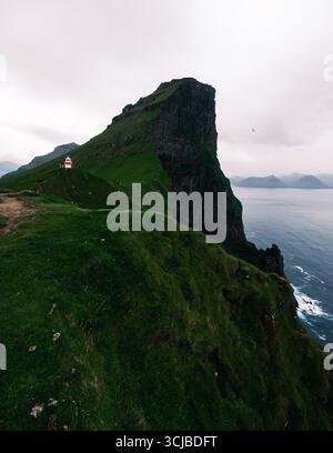 Una spettacolare scogliera si innalza bruscamente dalla costa delle Isole Faroe, adornata da lussureggiante erba verde e da un caratteristico faro. Le onde si infrangono contro la costa rocciosa sotto un cielo umido durante il crepuscolo. Foto Stock