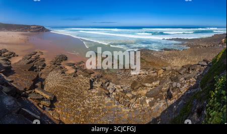 Una splendida scena costiera con onde turchesi che si infrangono su una riva rocciosa, incontrando una spiaggia sabbiosa sotto un vivace cielo blu a Praia das Maçãs Foto Stock