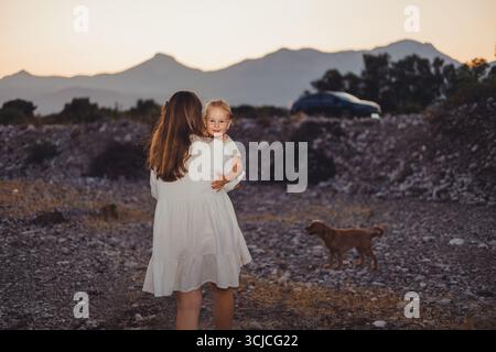 La vista posteriore della madre irriconoscibile in un abito bianco porta la figlia sorridente mentre cammina con un cane al tramonto. Lo sfondo panoramico delle montagne Foto Stock