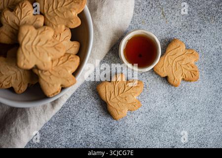Vista dall'alto dei biscotti a forma di foglia d'acero in una ciotola bianca su sfondo grigio. Una ciotola di sciroppo d'acero accompagna i biscotti, esaltandone il sapore autunnale Foto Stock