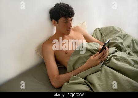 Un giovane sdraiato comodamente a letto, con uno smartphone. La luce del mattino illumina delicatamente la stanza, creando un'atmosfera rilassata mentre si impegna Foto Stock