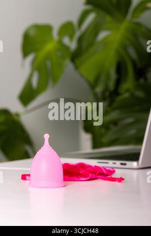 Una tazza mestruale rosa si trova su una scrivania bianca con un vivace panno rosa. Questa opzione ecocompatibile e sostenibile offre un'alternativa moderna alle mestruazioni Foto Stock