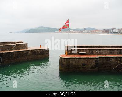 Una vibrante bandiera basca ondeggia orgogliosamente sulla costa di Zarautz, Paesi Baschi. Il mare sereno, la costa frastagliata e la pittoresca città creano un'atmosfera pittoresca Foto Stock