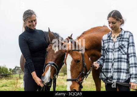 Una madre e la sua figlia adolescente legano mentre si occupano dei loro cavalli nella loro fattoria rurale in nuova Zelanda, mostrando equitazione naturale e sostegno Foto Stock