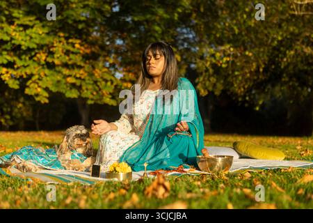 Una donna indiana siede pacificamente su una coperta da picnic, meditando in natura con il suo fedele cane al suo fianco. L'ambiente tranquillo è pieno di tranquillita' Foto Stock