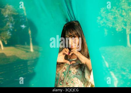 Una donna indiana sorridente in abbigliamento tradizionale si erge dietro il tessuto turchese, formando con le mani una forma a cuore in un parco soleggiato. La luce dorata enha Foto Stock
