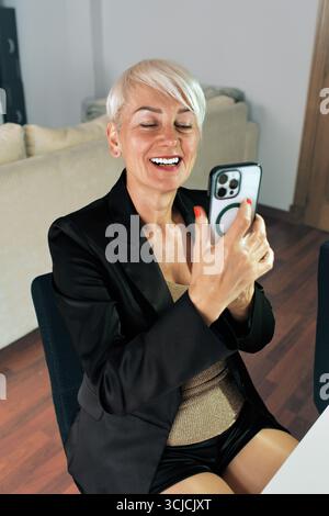 Donna con capelli corti biondi che sorride mentre lavora da casa utilizzando uno smartphone, vestita con un blazer formale, seduta comodamente in un moderno e ben illuminato Foto Stock