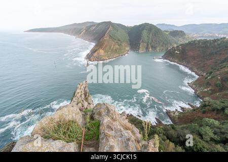 Una vista mozzafiato delle scogliere rocciose che conducono in una tranquilla baia sull'oceano, circondata da lussureggianti colline verdi e montagne lontane. Le onde si infrangono dolcemente Foto Stock