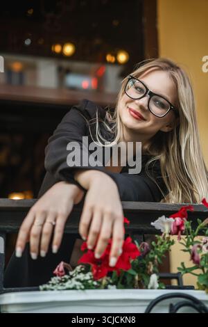 Una giovane donna bionda con gli occhiali sorride calorosamente mentre si appoggia su un binario adornato da fiori vivaci. Il suo abbigliamento casual e la sua espressione gioiosa trasudano Foto Stock