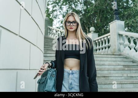 Giovane donna bionda che indossa una giacca nera e un paio di occhiali, in piedi con sicurezza all'aperto. Ha in mano una borsa blu e un telefono, con eleganti scale e verde Foto Stock