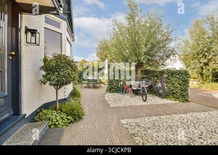 Splendida vista esterna con un affascinante passaggio pedonale, vegetazione lussureggiante e biciclette parcheggiate accanto a una pittoresca casa in mattoni bianchi con eleganti finestre Foto Stock