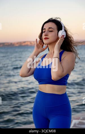 Una giovane donna in abbigliamento sportivo blu gode di un momento tranquillo ascoltando la musica all'aperto al tramonto, abbracciando l'armonia della natura e del fitness accanto al wa Foto Stock
