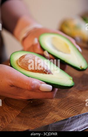Metà di avocado tagliate al momento tenute in mano su un tagliere di legno con verdure Foto Stock