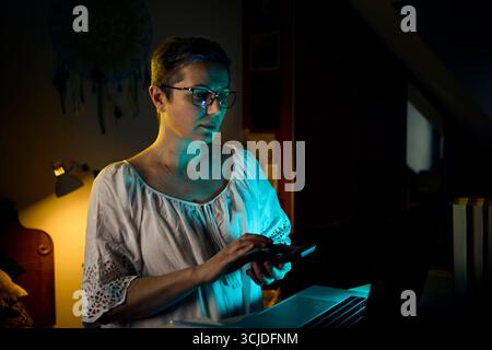 Una donna di mezza età con una camicetta bianca si indossa sul suo laptop mentre lavora da casa la sera. La calda lampada da scrivania mette in risalto la sua attenzione, rappresentativa Foto Stock