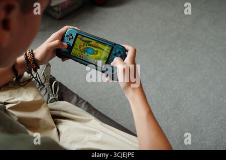 Adolescente con una pelle leggera che tiene in mano una console di gioco portatile che gioca a videogiochi, seduto in casa, concentrandosi intensamente sullo schermo, illustrando il concetto di dipendenza da gadget Foto Stock
