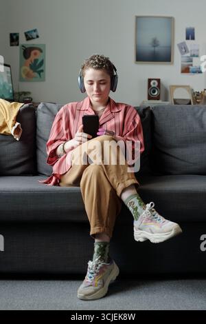 Ritratto di un adolescente caucasico seduto sul divano che indossa le cuffie con uno smartphone con un'espressione mirata che illustra la dipendenza da gadget in un ambiente domestico moderno Foto Stock