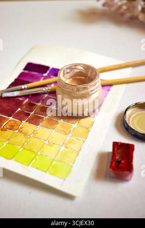 Piatti creativi con tavolozza di acquerelli, pennelli e vasetto di vernice su carta. Spazio di lavoro artistico con campioni vivaci in tonalità viola, giallo e rosso Foto Stock