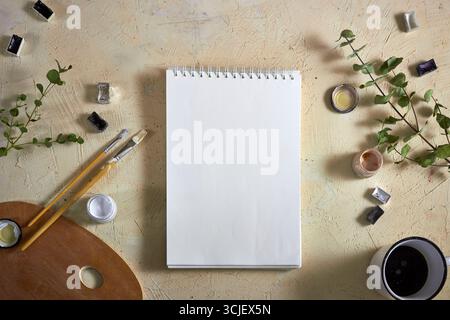 Vista dall'alto di uno schizzo vuoto circondato da colori ad acquerello, pennelli, tavolozza, vegetazione e una tazza di caffè. Sfondo creativo perfetto per lo spazio di lavoro Foto Stock