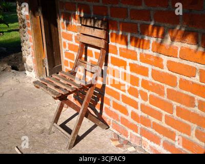 Una sedia di legno usurata appoggiata su una parete di mattoni arancioni Foto Stock