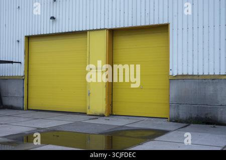 Due grandi banchine di carico di colore giallo brillante su un edificio industriale in metallo ondulato si riflettono in una pozzanghera sulla pavimentazione in cemento Foto Stock