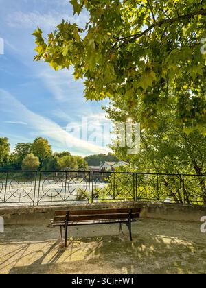 Panca in legno con vista sullo sbarramento des Forges, con ringhiera metallica, alberi e un vecchio edificio sullo sfondo, sotto la luce soffusa del sole Foto Stock