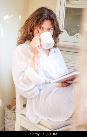 Una donna matura in abito bianco siede tranquillamente, sorseggiando un tè mentre legge un libro, circondata da un ambiente tranquillo che incarna eleganza e gioia, cr Foto Stock