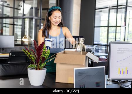 Donna asiatica che disimballa la scatola in un ufficio moderno, tiene una tazza di caffè e sorride Foto Stock