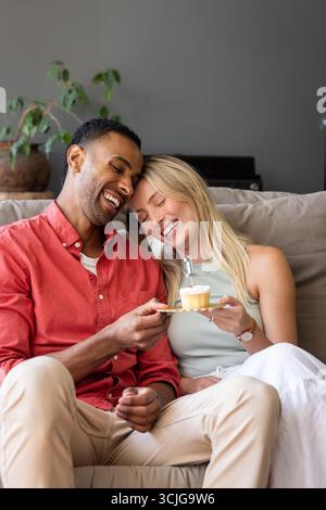 Coppia sul divano che condivide il cupcake, ridendo e godendosi un'accogliente festa a casa Foto Stock
