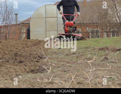 Un contadino in abiti scuri ara la terra sul suo terreno personale con un coltivatore di benzina di ferro rosso, un uomo allenta il terreno in un villaggio usando un aratro Foto Stock