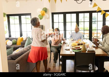 Donna anziana che porta la torta di compleanno mentre gli amici applaudono e festeggiano a casa Foto Stock