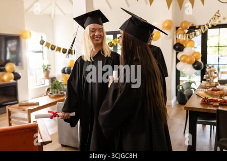 Laureati in berretti e camici che celebrano a casa con diplomi, sorridendo con gioia Foto Stock