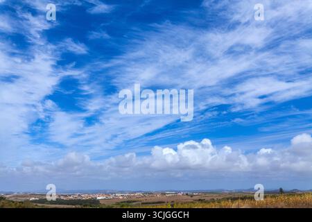 Un vasto paesaggio rurale sotto un cielo azzurro brillante con nuvole di neve, con campi dorati, colline lontane e un piccolo villaggio di Cambelas sull'horiz Foto Stock