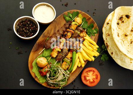 Piatto rustico in legno con grigliate miste mediorientali, tra cui kebab, kofta, shish tawook, patatine fritte, verdure fresche, focaccia e salsa tahini. Foto Stock