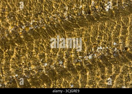 L'acqua increspata limpida crea motivi dorati su piccoli ciottoli. Foto Stock