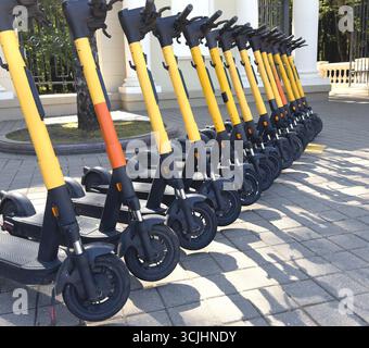 Fila di scooter elettrici gialli parcheggiati ordinatamente in un'area pavimentata della città, pronti per gli spostamenti urbani e i trasporti ecologici Foto Stock