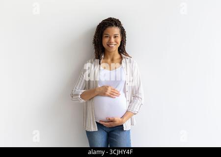 Sorridente donna nera incinta in piedi su sfondo bianco, tenendo la pancia con le mani Foto Stock