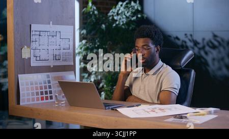 Uomo d'affari afroamericano architetto designer parlare telefono cellulare in ufficio uomo d'affari lavoro notturno lavoro online laptop cellulare chiamata maschio Foto Stock