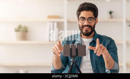 Influencer arabo maschile positivo che trasmette da casa, utilizzando smartphone e treppiede, seduto al tavolo in salotto Foto Stock