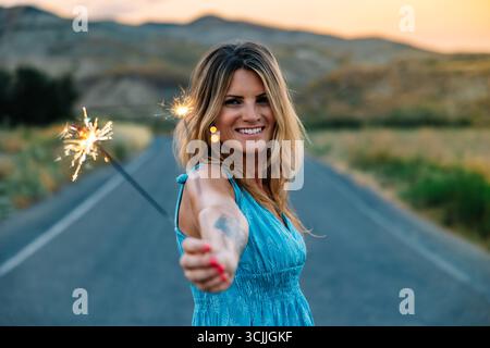 Donna che tiene sparkler sorridente al tramonto su una strada di campagna Foto Stock