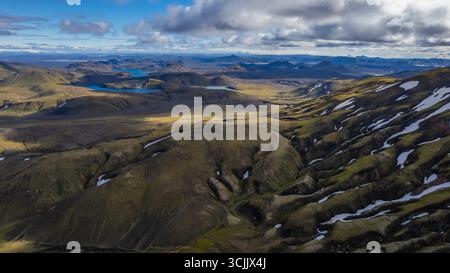 Vista aerea degli altopiani islandesi con colline vulcaniche ondulate macchie di neve e laghi blu sotto spettacolari nuvole in una remota natura selvaggia Foto Stock