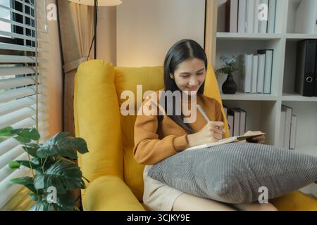 Una donna asiatica è seduta sul divano nel soggiorno. Sta tenendo un diario per registrare i ricordi e le esperienze passate. Tenere un diario è un piacere Foto Stock