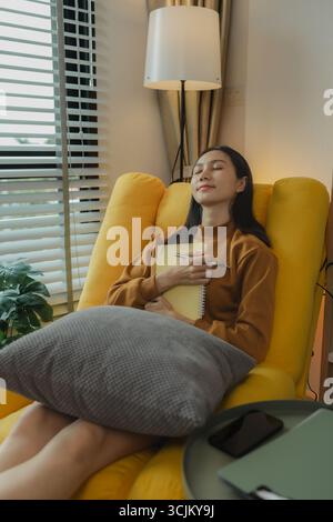 Una donna asiatica è seduta sul divano nel soggiorno. Sta tenendo un diario per registrare i ricordi e le esperienze passate. Tenere un diario è un piacere Foto Stock