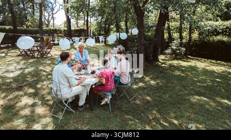 Vista aerea dei nonni, dei genitori e dei bambini che mangiano insieme all'esterno. Foto Stock