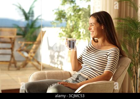 Una donna felice che beve un caffè seduto su una poltrona guarda lontano da una finestra di casa Foto Stock