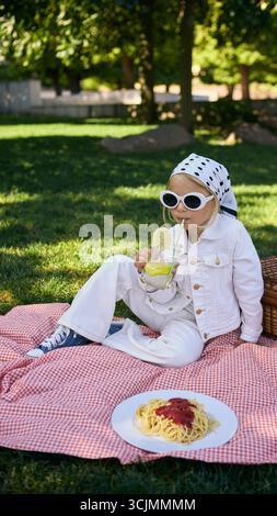 Ragazza con gli occhiali da sole sorseggiando limonata su coperta da picnic con piatto di spaghetti Foto Stock