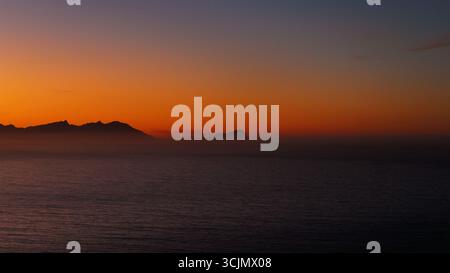 La vista aerea del tramonto infuocato dipinge il cielo sopra l'oceano scuro e tranquillo, sagomando le montagne lontane all'orizzonte. Città del Capo, Capo Occidentale, Sudafrica. Foto Stock