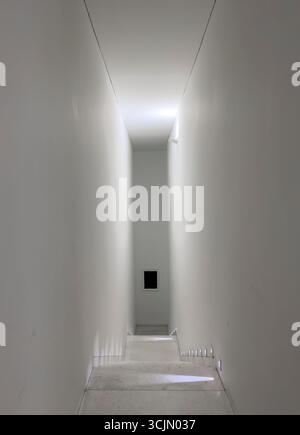 Scala interna minimalista con pareti bianche e illuminazione discreta che creano una prospettiva simile a un tunnel. Design architettonico pulito con passaggio stretto, Foto Stock