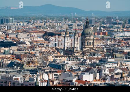Vista dello skyline della città con la basilica di Santo Stefano e una ruota panoramica che si staglia sullo sfondo di montagne lontane, Budapest, Ungheria. Foto Stock