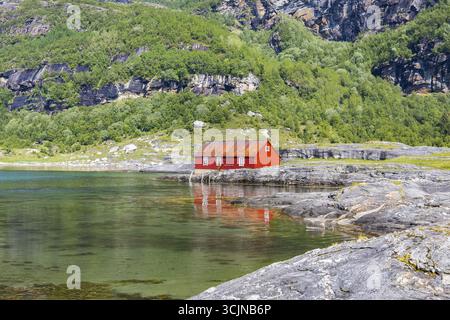 Vista di una vivace cabina rossa che si riflette sulle acque limpide e cristalline, annidata tra le coste rocciose e le lussureggianti montagne verdi di Mjelde, Bodo, Nordland, Norvegia. Foto Stock